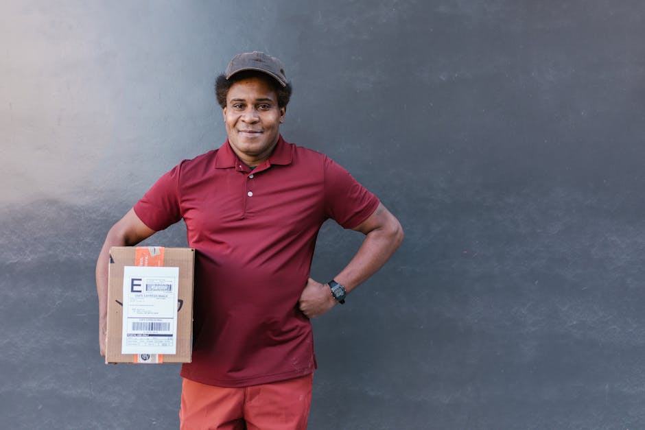 A delivery person with medium skin tone and short curly hair, wearing a maroon polo shirt, red trousers, and a dark grey cap, stands against a dark grey wall, smiling while holding a small cardboard p