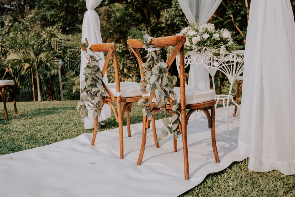 A pair of wooden chairs with rounded armrests and white cushions are decorated with cascading eucalyptus garlands and placed on a white fabric runner outdoors. Behind the chairs, there are white flora