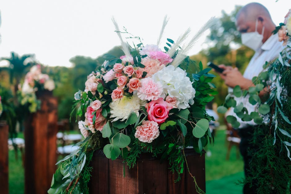 A vibrant floral arrangement featuring a mix of pale pink roses, white hydrangeas, and soft pink carnations, complemented by various green foliage and tall, wispy pampas grass. The bouquet is placed o